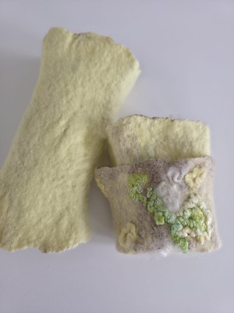 May include: Two pieces of felted wool. One is a long, pale yellow rectangular shape with a textured surface. The second piece is a shorter, light-colored rectangular cuff featuring intricate green and white embroidered floral details.