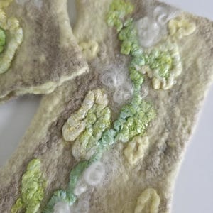 May include: Two rectangular felted wool pieces in shades of beige and cream, featuring raised, textured embroidery in light green, pale yellow, and white. The surface has a soft, fibrous appearance with organic, swirling patterns of stitched thread.