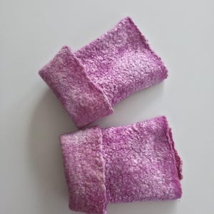 May include: Two handmade, felted wrist cuffs in shades of purple and white. The cuffs are rectangular with a folded-over cuff detail. The felted texture is visible, and the cuffs are displayed on a white background.