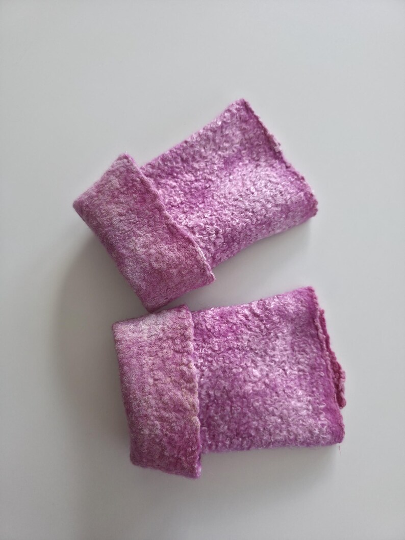 May include: Two handmade, felted wrist cuffs in shades of purple and white. The cuffs are rectangular with a folded-over cuff detail. The felted texture is visible, and the cuffs are displayed on a white background.
