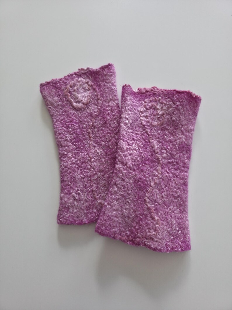 May include: A pair of handmade, fingerless gloves in a vibrant purple hue. The gloves feature a textured, felted wool design with a subtle floral embellishment near the wrist. The edges have a scalloped trim.