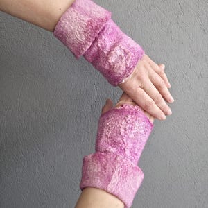 May include: A pair of pink felted wrist warmers. The wrist warmers are a vibrant pink with a textured surface and a folded cuff. A decorative floral element is visible on the top of each warmer. The background is a solid grey.