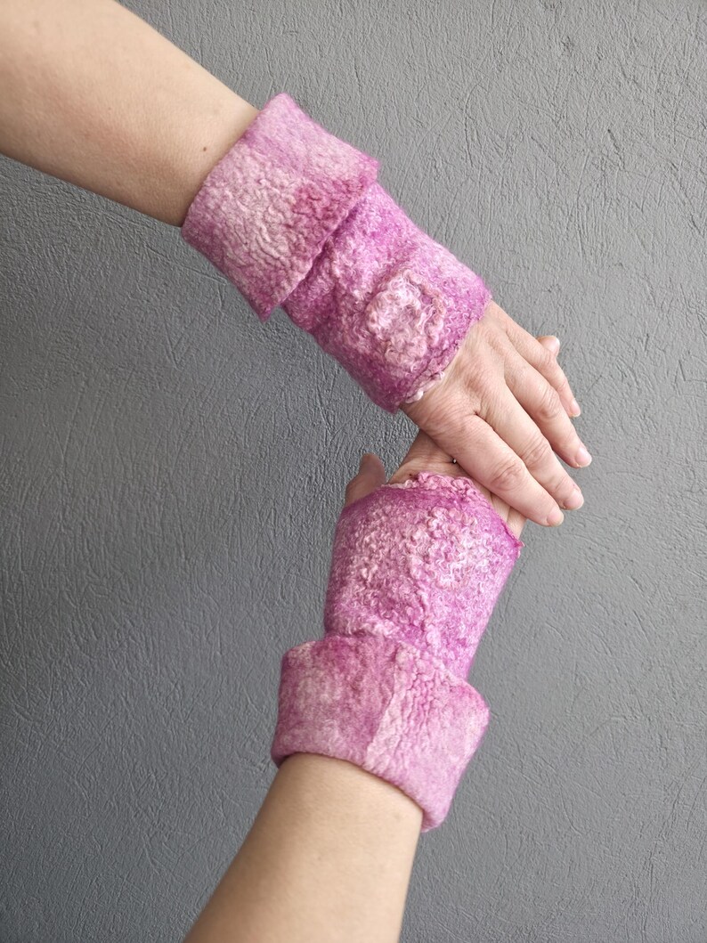 May include: A pair of pink felted wrist warmers. The wrist warmers are a vibrant pink with a textured surface and a folded cuff. A decorative floral element is visible on the top of each warmer. The background is a solid grey.
