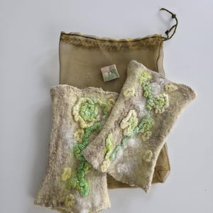May include: A pair of beige felted fingerless gloves featuring raised, textured floral patterns in shades of light green and white. The gloves are accompanied by a small, square, multi-colored felted piece and a sheer, olive-colored drawstring bag.