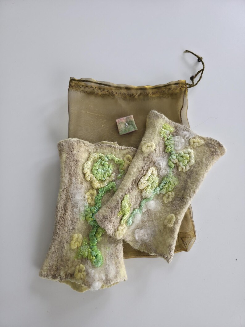 May include: A pair of beige felted fingerless gloves featuring raised, textured floral patterns in shades of light green and white. The gloves are accompanied by a small, square, multi-colored felted piece and a sheer, olive-colored drawstring bag.