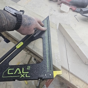 May include: A black CAL2 XL triangular layout tool with green markings and a yellow M-LOCK slider. The tool features a vertical scale ranging from 0 to 210, a yellow CUT TO C indicator, and is positioned on a piece of light-colored wood.