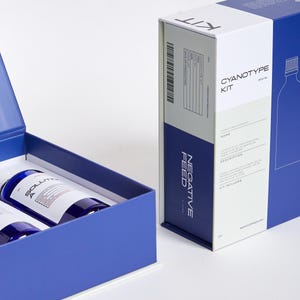 Pode incluir: Um kit de cianotipia com duas garrafas de solu&ccedil;&atilde;o dentro de uma caixa azul, ao lado de uma caixa maior com as palavras "Cyanotype Kit" e "Negative Feed" impressas. O kit inclui instru&ccedil;&otilde;es e componentes para criar impress&otilde;es de cianotipia.