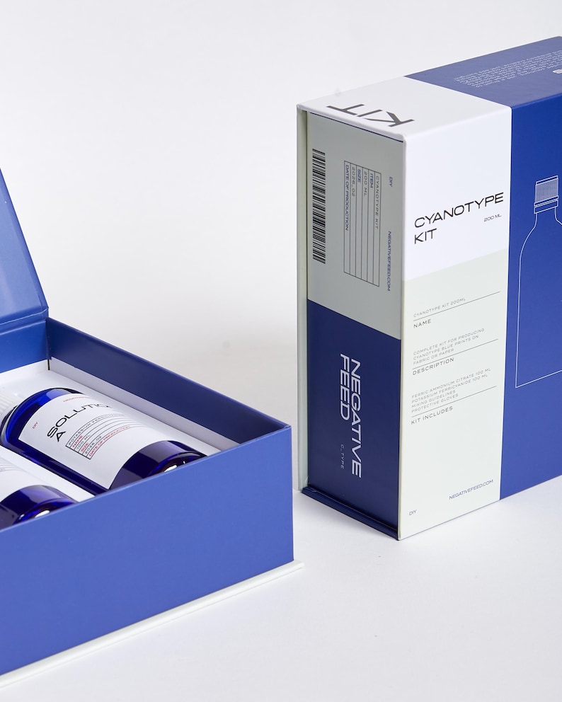 Pode incluir: Um kit de cianotipia com duas garrafas de solu&ccedil;&atilde;o dentro de uma caixa azul, ao lado de uma caixa maior com as palavras "Cyanotype Kit" e "Negative Feed" impressas. O kit inclui instru&ccedil;&otilde;es e componentes para criar impress&otilde;es de cianotipia.