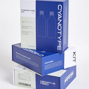 Op de afbeelding: Gestapelde cyanotype kitdozen in wit en blauw. De dozen tonen de woorden "CYANOTYPE KIT" en "NEGATIVE FEED" in het wit. Eén doos heeft een diagram van twee flessen. De dozen zijn op een witte achtergrond geplaatst.