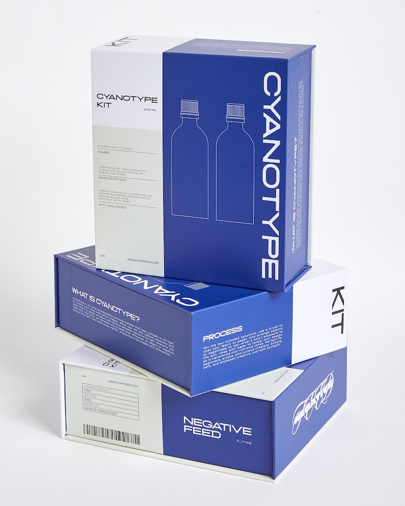 Pode incluir: Caixas de kit de cianotipia empilhadas em branco e azul. As caixas apresentam as palavras "CYANOTYPE KIT" e "NEGATIVE FEED" em branco. Uma caixa tem um diagrama de duas garrafas. As caixas est&atilde;o dispostas sobre um fundo branco.