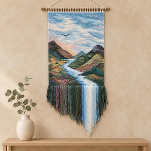 May include: A rectangular woven wall hanging featuring a landscape scene with mountains, a winding river, and a bird in a blue sky. The bottom edge has long, multi-colored yarn fringe in shades of green, purple, blue, and white hanging down.
