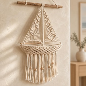 May include: A cream-coloured macrame wall hanging shaped like a sailboat, suspended from a horizontal wooden dowel. The piece features intricate knotted patterns forming the sails and hull, with long fringe at the bottom accented by wooden beads.