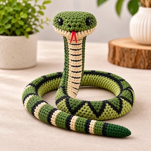May include: A crocheted snake toy featuring a green, black, and cream-coloured patterned body. The snake has a wide head with black plastic eyes and a protruding red yarn tongue. The underside of the neck and body displays a cream-coloured ribbed texture.