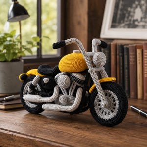 Puede incluir: Una motocicleta de ganchillo hecha a mano en amarillo, negro y gris. La motocicleta de juguete tiene un depósito de combustible amarillo, un asiento negro y ruedas grises. El manillar y los detalles del motor también son grises. La motocicleta se exhibe sobre una superficie de madera.