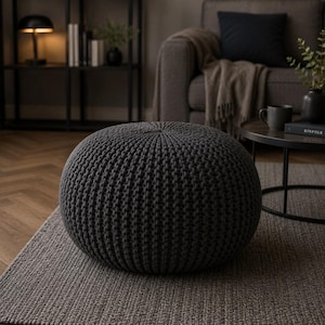 May include: A round, dark grey pouf featuring a thick, textured knit construction. The ottoman has a dense, uniform weave pattern that creates a rounded, firm shape, providing a solid surface for seating or resting feet in a living space.