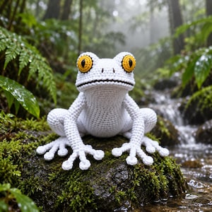 Könnte beinhalten: Eine weiße, gehäkelte Froschfigur mit großen, markanten Augen, leuchtend gelben Iris und dunklen Pupillen. Das Wesen hat eine texturierte, gestrickte Oberfläche, ein breites Maul mit einer dunklen horizontalen Linie und vier gespreizte Füße.