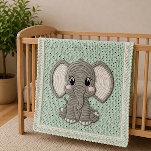 Puede incluir: Una manta rectangular de ganchillo en verde menta claro con una aplicación central de elefante gris con grandes orejas blancas, mejillas rosadas y ojos negros. La manta tiene un patrón de punto texturizado y un borde crema festoneado.