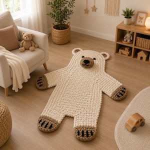 May include: A cream-colored, hand-crocheted bear-shaped rug featuring a dark brown snout, black eyes, and brown paw pads with black stitched details. The rug is crafted from thick yarn and is laid flat on a light-colored wooden floor surface.