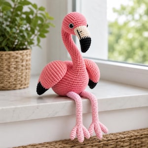 May include: A handmade pink crochet flamingo with black accents on the wings and beak. The flamingo has black eyes and is sitting with its legs dangling. The flamingo is a decorative item.