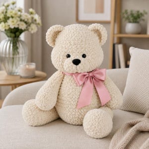 May include: A cream-colored plush teddy bear made of soft, textured yarn. The bear features round ears, black plastic eyes, and a black stitched nose. A wide, smooth, dusty pink satin ribbon is tied into a bow around the bear's neck.