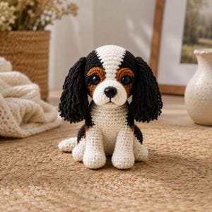 Könnte beinhalten: Ein gehäkelter Cavalier King Charles Spaniel Hund mit einem Farbschema aus Schwarz, Weiß und Braun. Das handgefertigte Plüschtier hat einen weißen Körper, schwarze Ohren und braune Akzente um die Augen. Es sitzt auf einer gewebten Matte.