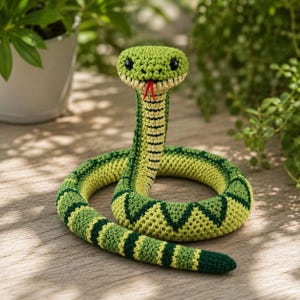 May include: A crocheted snake toy featuring a vibrant green and yellow patterned body with dark green diamond accents. The snake has a raised head with black bead eyes and a small red yarn tongue protruding from its mouth, coiled in a circle.