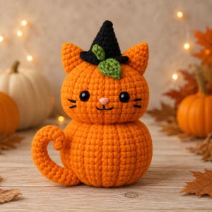 May include: A crocheted orange pumpkin cat figurine featuring a black witch hat with a small green leaf accent. The cat has black embroidered whiskers, a pink nose, and two black eyes. The body is round and textured with a curled tail on the side.