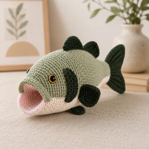May include: A crocheted fish toy featuring a sage green body, a pale pink open mouth, and dark forest green fins. The fish has a round, black eye with a gold-coloured iris. The texture is created with tight, uniform yarn stitches throughout.
