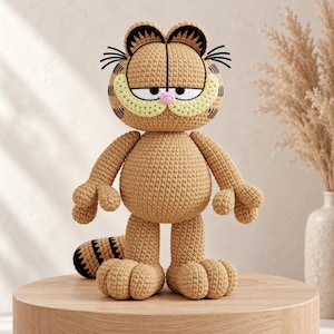 Può includere: Un peluche all'uncinetto color marrone chiaro che raffigura un gatto dei cartoni animati con una grande testa rotonda, occhi neri ricamati, un piccolo naso rosa e un ampio muso giallo chiaro. La figura presenta strisce marrone scuro sulla testa.