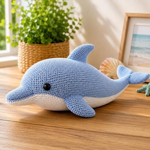 May include: A light blue and white crocheted dolphin plush toy with a smooth, rounded snout, a prominent dorsal fin, and a dark, round plastic eye. The body features a textured yarn stitch pattern, transitioning from a blue top to a white belly.
