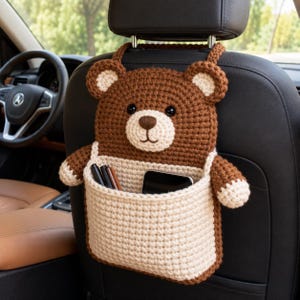 May include: A brown and cream crocheted bear-shaped car seat organiser hanging from a headrest. The bear features a dark brown body, cream-coloured muzzle and inner ears, and a large cream pocket containing several pens and a black smartphone.