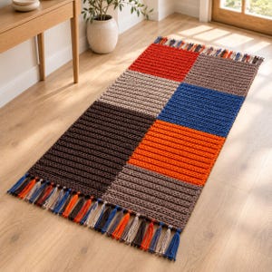 May include: A rectangular crochet rug featuring a colour-blocked pattern of red, brown, blue, orange, and beige squares. The rug has a ribbed texture and is finished with multi-coloured yarn tassels along the two shorter ends of the piece.