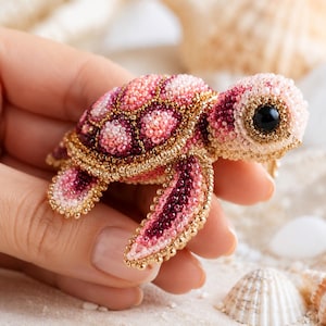 May include: A beaded turtle brooch crafted with shades of pink, magenta, and cream seed beads. The turtle features a prominent black bead eye, a detailed shell pattern, and gold-toned metallic bead edging along the fins and the perimeter of the shell.