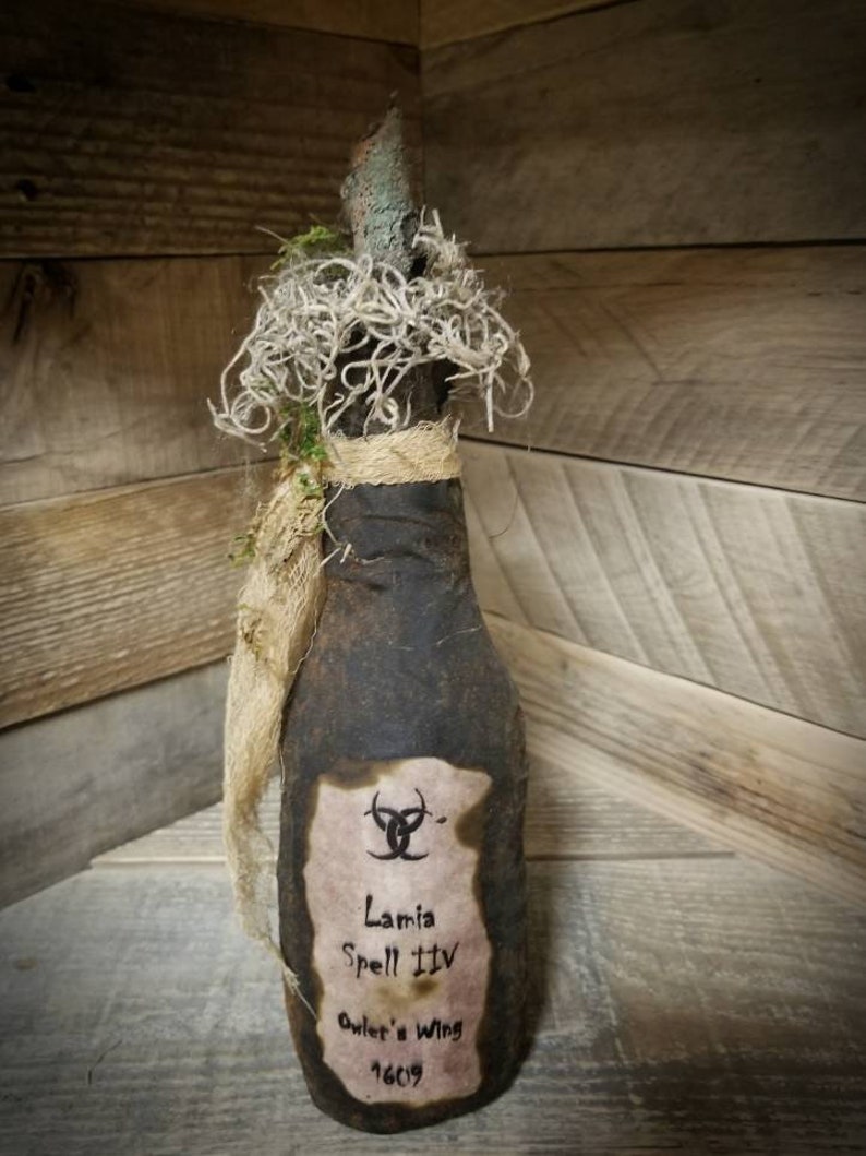 Op de afbeelding: Een donkerbruine glazen fles met een kurkstop en een etiket met de tekst "Lamia Spell IIIV Owl's Wing 1609". De fles is versierd met touw en mos.
