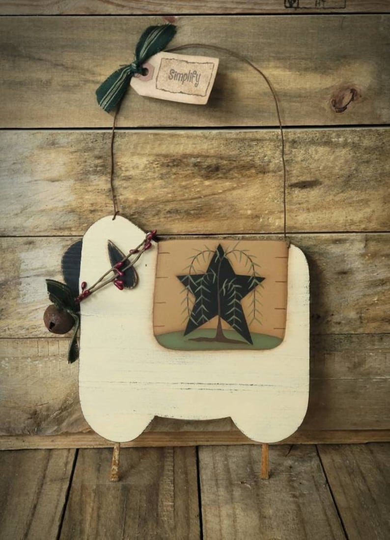 Puede incluir: Un colgante de pared r&uacute;stico de madera color crema con un dise&ntilde;o de estrella y &aacute;rbol. La estrella es negra con un borde verde, y el fondo es marr&oacute;n claro. Una etiqueta con la palabra "Simplify" cuelga de una cinta verde. Una peque&ntilde;a campana y bayas a&ntilde;aden detalles.