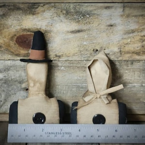 May include: Two primitive fabric dolls with black buttons and simple features. The doll on the left is wearing a black hat with a brown band. The doll on the right is wearing a white bonnet. Both dolls are standing on a wooden surface.