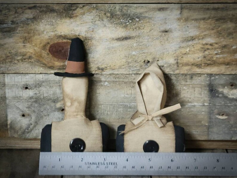 May include: Two primitive fabric dolls with black buttons and simple features. The doll on the left is wearing a black hat with a brown band. The doll on the right is wearing a white bonnet. Both dolls are standing on a wooden surface.