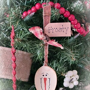 May include: A wooden spoon painted to look like a snowman with a red nose and black eyes. The spoon is hanging from a Christmas tree with red beads and a tag that says "Frosty".