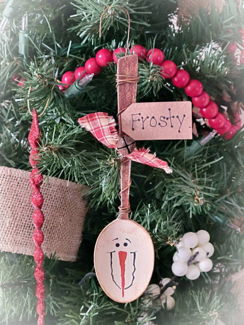 May include: A wooden spoon painted to look like a snowman with a red nose and black eyes. The spoon is hanging from a Christmas tree with red beads and a tag that says "Frosty".
