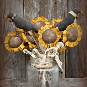 May include: A rustic arrangement of faux sunflowers and black birds in a clear glass jar. The sunflowers have brown centers and yellow petals. The birds have black bodies and white accents. The jar has a textured surface and is sitting on a wooden surface.