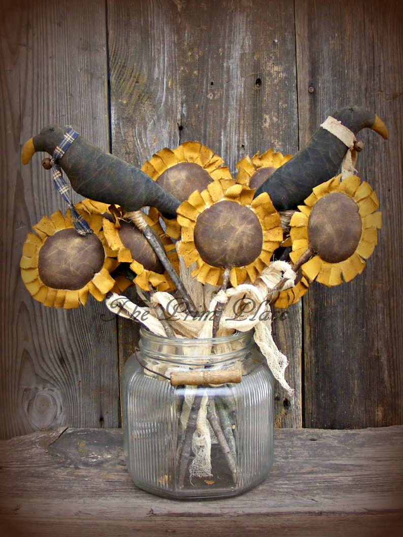 May include: A rustic arrangement of faux sunflowers and black birds in a clear glass jar. The sunflowers have brown centers and yellow petals. The birds have black bodies and white accents. The jar has a textured surface and is sitting on a wooden surface.