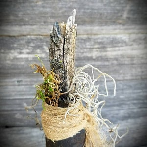 Op de afbeelding: Een rustieke, handgemaakte decoratieve fles met een natuurlijke houten bovenkant, gewikkeld in jute en versierd met gedroogd mos en wijnstokken. De fles is een uniek en charmant stuk voor thuisdecoratie.