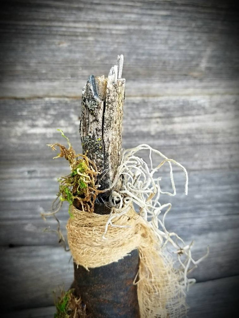 Op de afbeelding: Een rustieke, handgemaakte decoratieve fles met een natuurlijke houten bovenkant, gewikkeld in jute en versierd met gedroogd mos en wijnstokken. De fles is een uniek en charmant stuk voor thuisdecoratie.