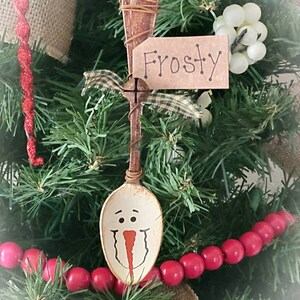 May include: A wooden spoon painted to look like a snowman with a red nose and black eyes. The spoon is hanging on a Christmas tree with a tag that says "Frosty".