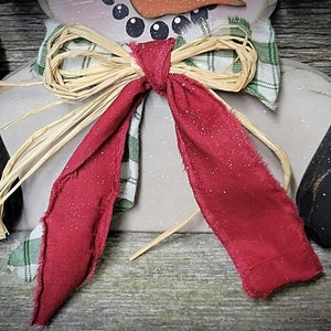 May include: A red and green plaid bow with a straw bow on top. The bow is tied around the neck of a snowman.