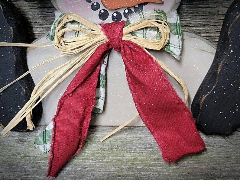 May include: A red and green plaid bow with a straw bow on top. The bow is tied around the neck of a snowman.