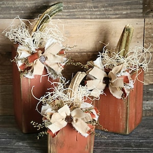 May include: Three rustic wooden pumpkins decorated with burlap, twine, and dried flowers. The pumpkins are painted in a distressed orange color and have a vintage farmhouse style.