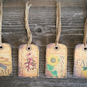 May include: Four autumn-themed gift tags with twine. The tags feature images of corn, leaves, sunflowers, and pumpkins. The text on the tags reads "Harvest Time", "Falling Leaves", "Nature Walks", and "Hello Fall".