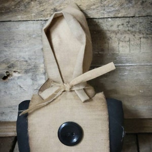 May include: A handmade fabric doll with a black button for an eye. The doll is wearing a brown fabric hood and a black fabric skirt.