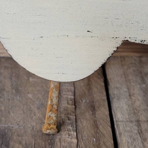 Puede incluir: Primer plano de un peque&ntilde;o objeto de madera blanquecina con un borde curvo y una sola pata de metal oxidado. La madera tiene un aspecto envejecido y desgastado con vetas visibles y detalles negros. La pata es rectangular y muestra signos de &oacute;xido.